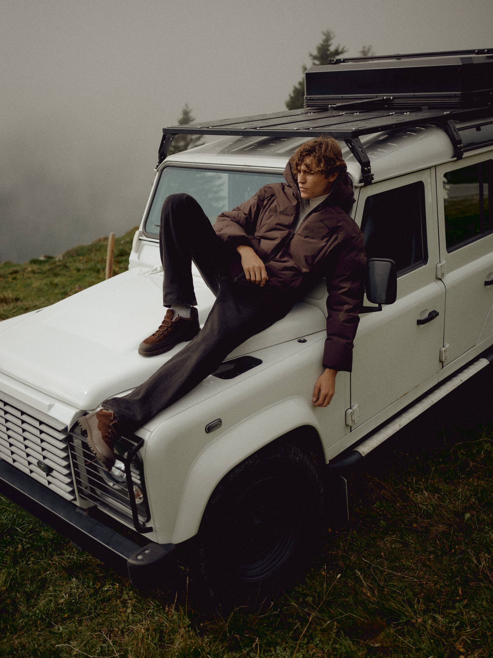 Man wearing a cocoa bean CASPARA Boreal puffer jacket outdoors on the hood of an off-road vehicle.